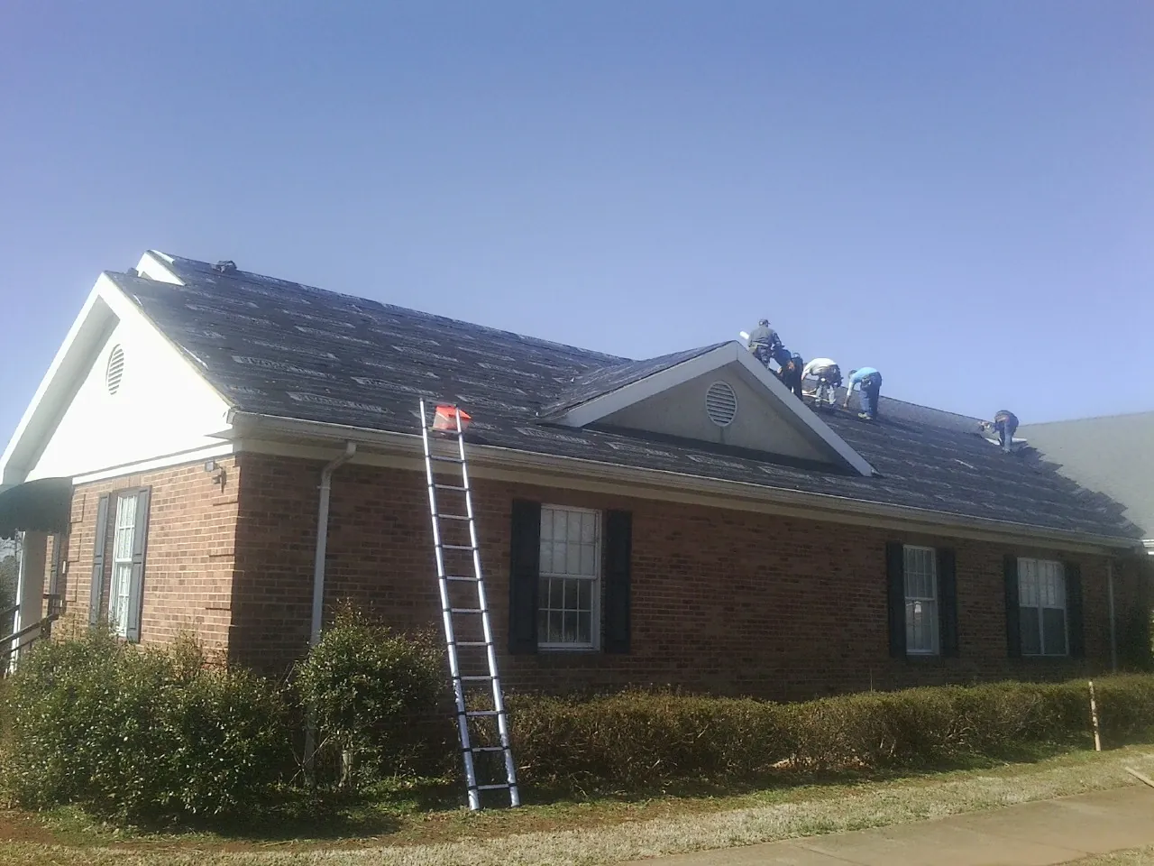 Crew tearing shingles off a brick ranch roof with a ladder set — Emergency Roof Repair work by our crew in Six Mile